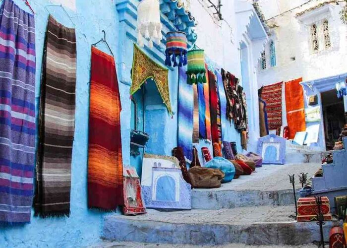 Colorful textiles hanging along the blue-washed walls of Chefchaouen, Morocco, on a cultural stop during the Marrakech to Chefchaouen Desert Tour.