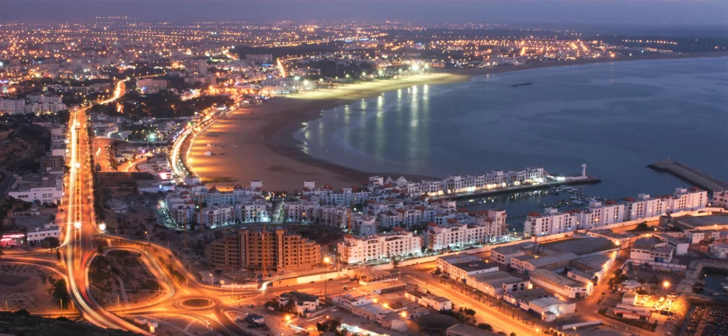 Night view of Agadir's glowing cityscape with illuminated coastline, marina, and beach in Morocco.