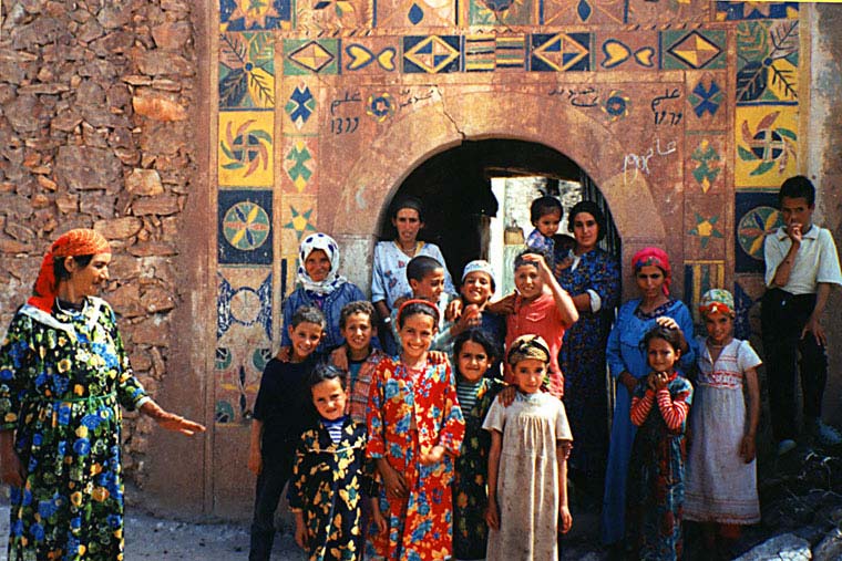 A Berber family smiles together in traditional attire in their Atlas Mountains village, with colorful tribal patterns adorning their home's entrance.