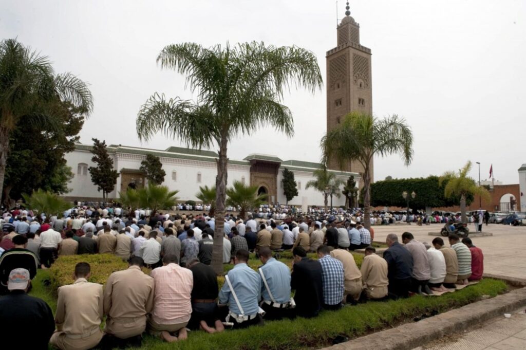 Moroccan community performing Friday prayers outdoors with a minaret in the background."