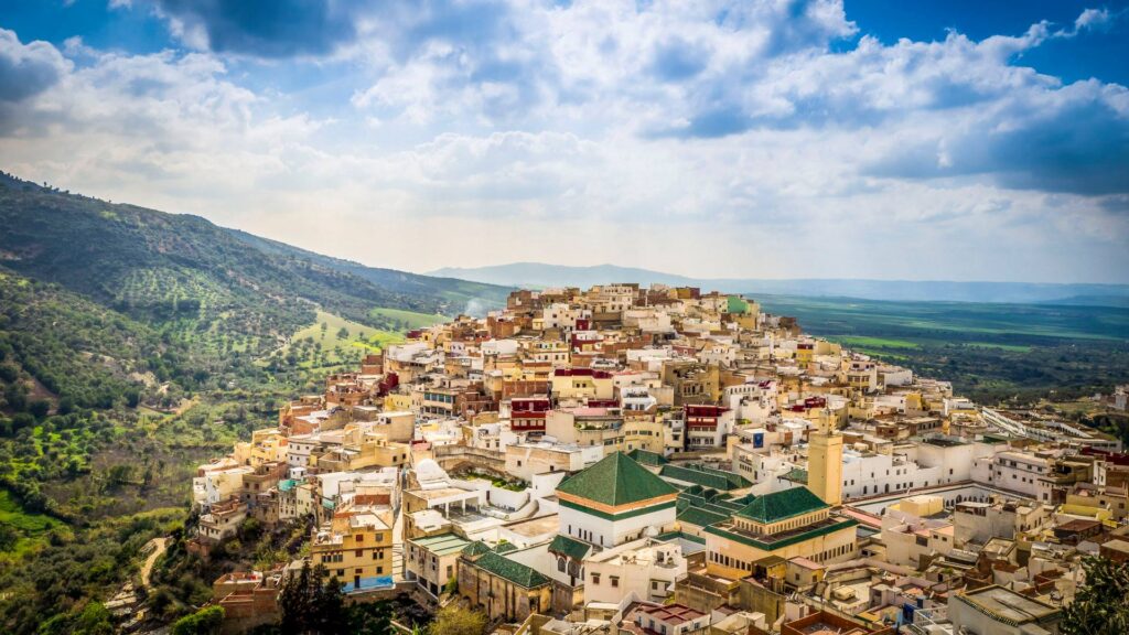 Panoramic view of a densely packed hilltop town in Morocco with surrounding green landscapes under a cloudy sky.