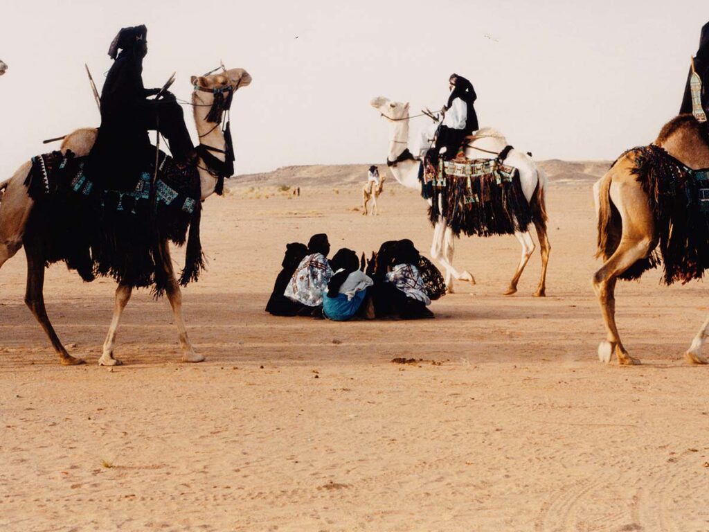 Tuareg camel riders and local tribespeople in traditional attire gathered in the Sahara Desert, demonstrating their adaptation to desert life.