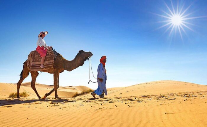 A tourist riding a camel led by a guide through the sandy dunes under the bright sun in the Moroccan desert
